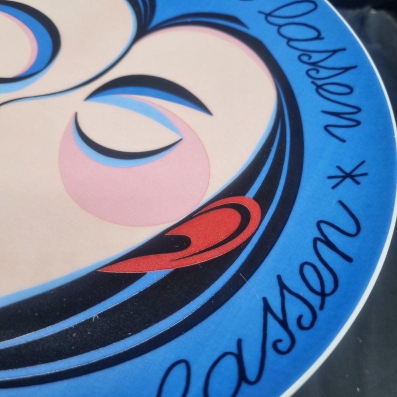 May include: A decorative plate with a stylized illustration of a woman's face. The design features a light pink face, black hair, and a blue border with the name "Lassen" in cursive. The plate has a red accent and a white rim.