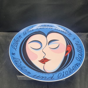 May include: A decorative plate with a blue rim and a central design featuring two faces in profile, their lips touching. The faces are in shades of pink and black, with red lips and a red ear. The plate has text in cursive around the edge.