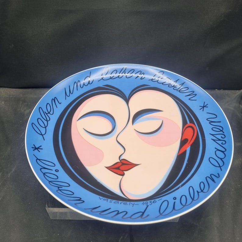 May include: A decorative plate with a blue rim and a central design featuring two faces in profile, their lips touching. The faces are in shades of pink and black, with red lips and a red ear. The plate has text in cursive around the edge.