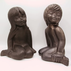 May include: Two ceramic figurines of children sitting with their legs crossed. The figurines are a dark brown colour and have a smooth, matte finish.