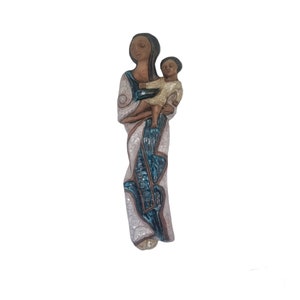 Op de afbeelding: Een keramische wandsculptuur van een moeder en kind. De moeder draagt een blauwe en witte gepatroneerde jurk en houdt het kind in haar armen. Het kind draagt een witte jurk. De sculptuur is in moderne stijl.