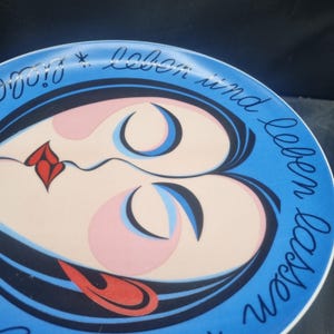 May include: A vintage ceramic plate with a blue rim and a central design featuring a stylized face with pink and blue accents. The plate has the German text "Leben und leben lassen" in a cursive font around the edge.