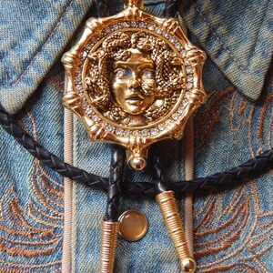May include: Gold-toned bolo tie featuring a Medusa pendant with clear crystal accents. The pendant is set on a black braided cord with gold-colored metal tips and a circular clasp. The tie is displayed on a denim shirt.
