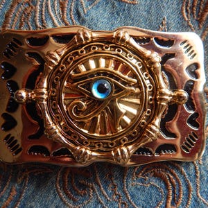 May include: A rectangular gold-toned belt buckle featuring a central circular emblem with a raised Eye of Horus design. The eye has a bright blue cabochon centre. The buckle has black enamel accents and a decorative border with heart-shaped cutouts.