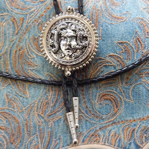 Può includere: Un bolo tie argentato con un ciondolo rotondo che presenta un'immagine dettagliata del viso di Medusa. Il ciondolo è circondato da un bordo di perline. Il bolo tie è su uno sfondo di denim blu con un motivo ricamato.