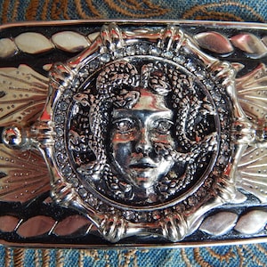 May include: A silver belt buckle with a black background. The buckle features a detailed design of a woman's face surrounded by snakes, with rhinestones around the edge.