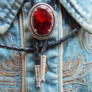 Peut inclure: Un bolo tie avec un pendentif en argent et pierre rouge sur une chemise en jean avec des détails brodés.