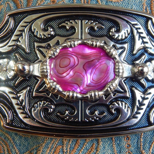 Könnte beinhalten: Rechteckige Gürtelschnalle mit silbernem und schwarzem Filigranmuster. In der Mitte befindet sich eine ovale, rosa Abalone-Muschel. Florale und scrollartige Details zieren die Ränder und schaffen ein dekoratives Accessoire.