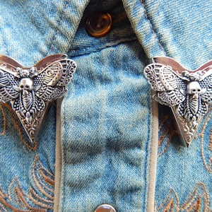 May include: Two silver-toned moth-shaped collar pins with a skull design. The pins are attached to a blue denim shirt.
