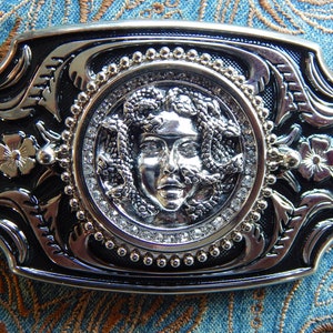 Peut inclure: Une boucle de ceinture argentée avec un fond noir. La boucle présente un motif détaillé d'un visage de femme avec des serpents dans les cheveux. Le visage est entouré d'un cercle de strass.