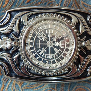 Może przedstawiać: Srebrna klamra do paska z czarnym tłem. Klamra ma okrągły wzór z kompasem i runami wokół krawędzi. Klamra ma po obu stronach ozdobny wzór kwiatowy.