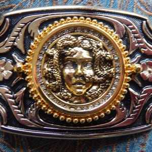 Pode incluir: Fivela de cinto retangular com uma cabeça de Medusa dourada no centro, rodeada por um círculo de strass transparentes e contas douradas. A fivela tem um fundo preto com desenhos de filigrana dourados e prateados e pequenas flores douradas.