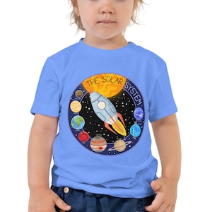 May include: A light blue short-sleeved t-shirt featuring a circular graphic of the solar system with a central rocket ship, the sun, and various planets orbiting against a black starry background with the text THE SOLAR SYSTEM printed above.