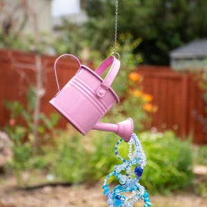 May include: A small, pink metal watering can hangs from a chain, appearing to pour out a stream of blue and clear beads. The beads are a mix of shapes and sizes, creating a decorative, whimsical effect. The background is a blurred outdoor scene.