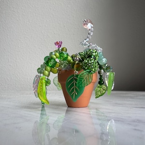 May include: A decorative beaded plant in a small terracotta pot. The plant features green and clear beads arranged to resemble leaves and stems, with a few pink and clear accents. The pot sits on a reflective surface.