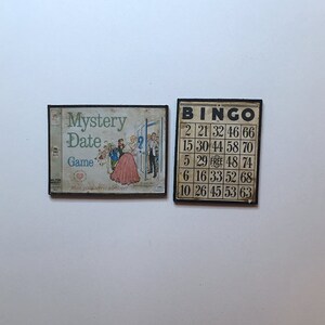 Puede incluir: Dos tableros de juego vintage. El primer tablero es de color marrón claro con una imagen de una pareja bailando y el texto "Mystery Date Game". El segundo tablero es negro con texto blanco que dice "BINGO" y una cuadrícula de números.