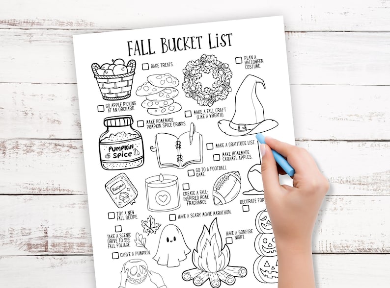 May include: Black and white Fall Bucket List printable with illustrations of fall activities. Includes text such as "Pumpkin Spice", "Bonfire Night", and "Carve a Pumpkin". A hand with a blue pen is partially visible, marking a box.