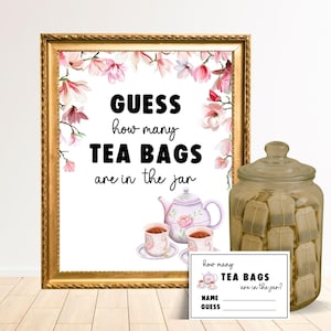 May include: A gold-framed sign reads "GUESS how many TEA BAGS are in the jar". A glass jar filled with tea bags and a small card with the same text are also present. The sign has a floral border with pink flowers.