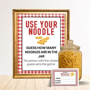 May include: A framed game sign with the text "USE YOUR NOODLE" and "GUESS HOW MANY NOODLES ARE IN THE JAR". A glass jar filled with uncooked pasta is next to the sign, along with a small game card.
