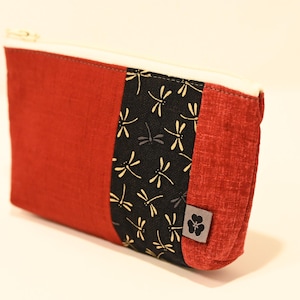 Può includere: Una pochette con cerniera rossa e nera con un motivo a libellule. La pochette ha una piccola etichetta nera con un disegno di un fiore.