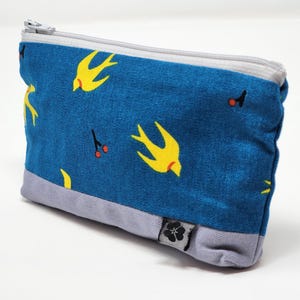 Peut inclure: Une pochette en tissu bleu avec un motif répété d'oiseaux jaunes et de petites baies rouges. La pochette a une base grise et une fermeture éclair argentée. Une petite étiquette noire et blanche est attachée en bas.