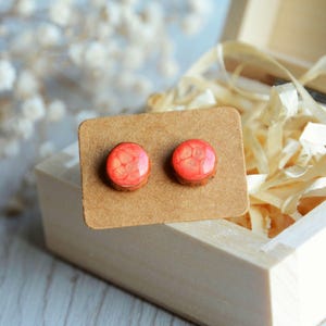 May include: A pair of round stud earrings with a red and orange pattern, set on a brown cardboard backing. The earrings have a wooden base and a glossy, resin-like top. The earrings are displayed inside a wooden box with decorative paper.