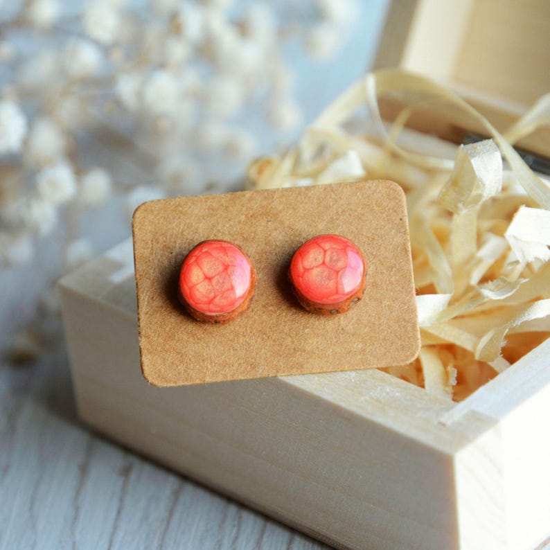 May include: A pair of round stud earrings with a red and orange pattern, set on a brown cardboard backing. The earrings have a wooden base and a glossy, resin-like top. The earrings are displayed inside a wooden box with decorative paper.
