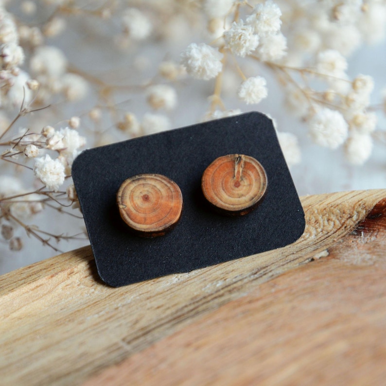May include: A pair of wooden earrings with a natural wood grain pattern. The earrings are round and have a light brown color with darker brown rings.