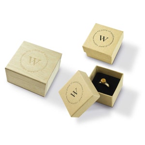 May include: Three jewelry boxes, two in light brown cardboard and one in light wood. The boxes are open, revealing a ring with a gold-colored setting and a black interior. The boxes have the text "my piece of wood wooden jewellery" and a "W" logo.