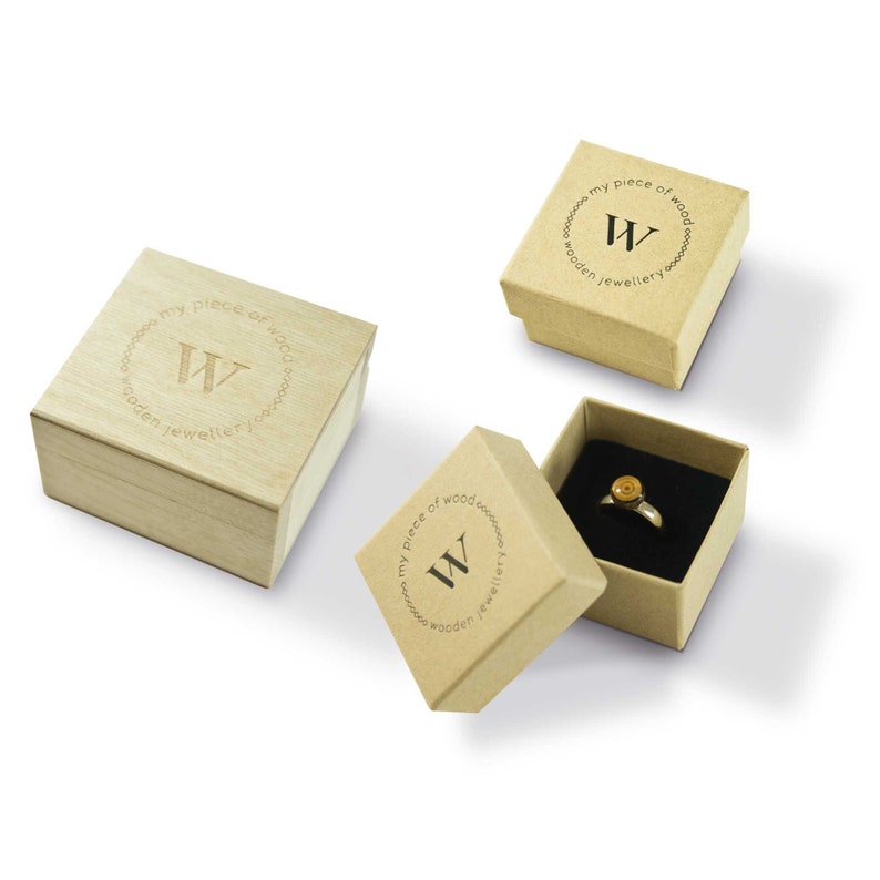 May include: Three jewelry boxes, two in light brown cardboard and one in light wood. The boxes are open, revealing a ring with a gold-colored setting and a black interior. The boxes have the text "my piece of wood wooden jewellery" and a "W" logo.