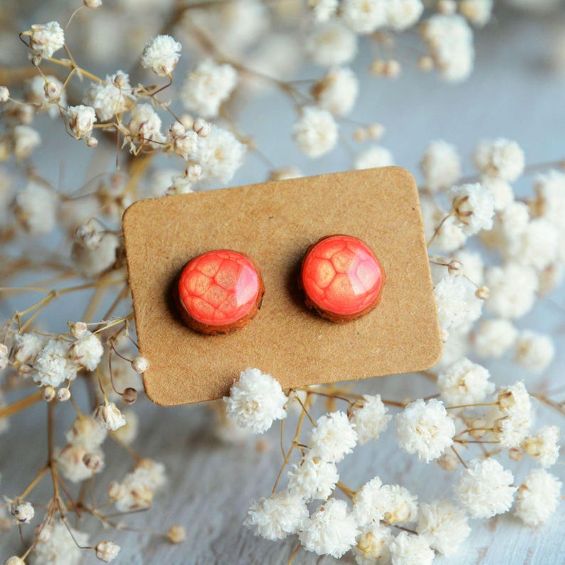 May include: A pair of round stud earrings with a reddish-orange, textured surface. The earrings are set on a brown wooden base and displayed on a brown card. The background features white flowers.