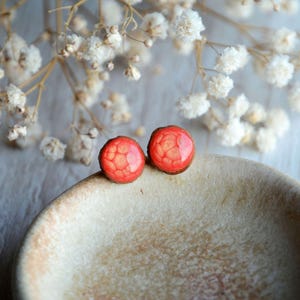 May include: A pair of round, red stud earrings with a textured, geometric pattern. The earrings are set on a light brown base and rest on a beige, ceramic surface. Delicate white flowers are in the background.