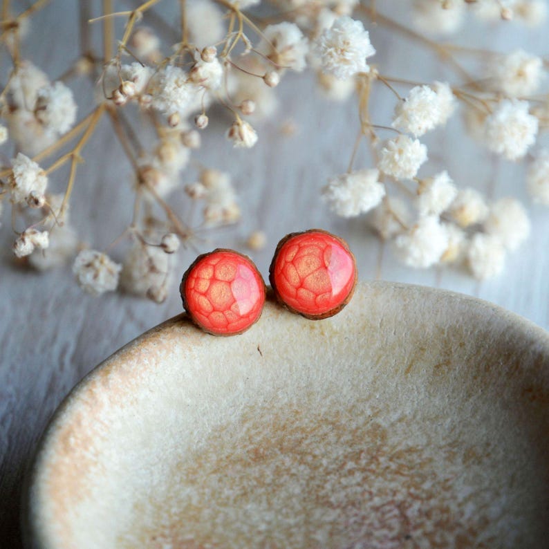 May include: A pair of round, red stud earrings with a textured, geometric pattern. The earrings are set on a light brown base and rest on a beige, ceramic surface. Delicate white flowers are in the background.