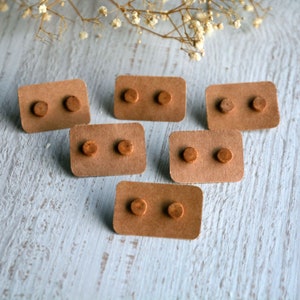 K&ouml;nnte beinhalten: Sechs Paar Holzstecker-Ohrringe, pr&auml;sentiert auf kleinen, rechteckigen Pappkarten. Die Ohrringe sind klein, rund und hellbraun. Die Karten sind hellbraun und auf einer hellblau-wei&szlig;en Oberfl&auml;che angeordnet.