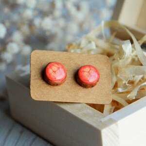 May include: A pair of round stud earrings with a red, textured top and a brown wooden base. The earrings are displayed on a small, brown card inside a wooden box with wood shavings.