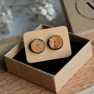 May include: A pair of wooden earrings in a small cardboard box. The earrings are round and have a natural wood grain pattern. The box is brown and has the word "wood" printed on it.