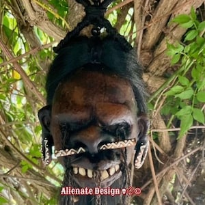 Può includere: Una maschera marrone stilizzata con capelli neri e dettagli facciali intricati, tra cui un ampio sorriso e decorazioni di perline. La maschera è sospesa su uno sfondo di rami di alberi e fogliame. Il testo "Alienate Design" è visibile.