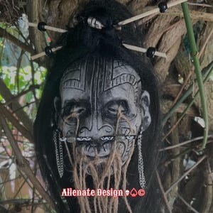 May include: A shrunken head replica with grey skin, intricate black facial markings, and long black hair styled with wooden sticks and beads. The face features twine hanging from the eyes and mouth, silver chains, and the text Alienate Design.