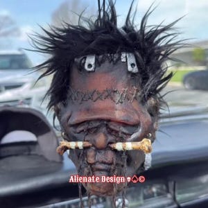 May include: A handcrafted, macabre decorative head with stitched details, metal accents, and a chain. The head features a dark complexion, black hair, and bone-like elements. The text "Alienate Design" is visible.