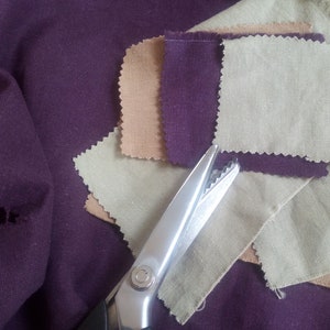 May include: A close-up of a pair of pinking shears on a purple fabric background. The shears are open and the blades are visible. There are four fabric swatches in the background, one each of brown, purple, beige, and light green.