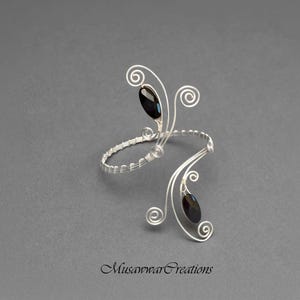 May include: A silver-toned arm cuff with two black oval-shaped gemstones. The cuff features a wire-wrapped design with decorative swirls and is set against a gray background. The text "MusawwarCreations" is at the bottom.