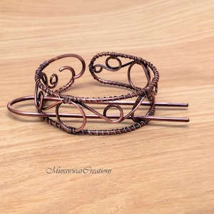 Op de afbeelding: Een haarstick van koperdraad met een decoratief ontwerp. De stick is gebogen in een cirkel met een kleinere cirkel in het midden. De stick is ook versierd met een swirl-ontwerp.