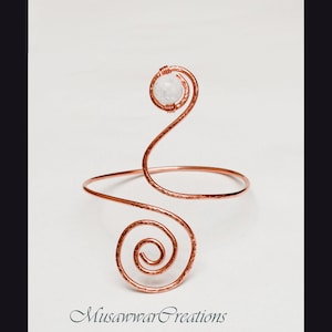 Puede incluir: Brazalete de brazo color cobre con textura martillada y diseño en espiral. Una piedra redonda y transparente está engastada en la parte superior del brazalete. El brazalete está diseñado para ser usado en la parte superior del brazo. El texto "Musawwar Creations" está en la parte inferior.