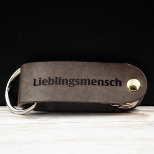 May include: A brown leather keychain with a silver ring and a gold button. The keychain is engraved with the text "Lieblingsmensch".