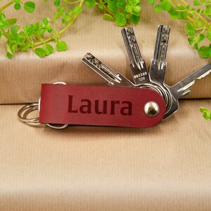 May include: Red leather keychain with the name "Laura" engraved on it. The keychain has a silver ring and is attached to a set of keys.