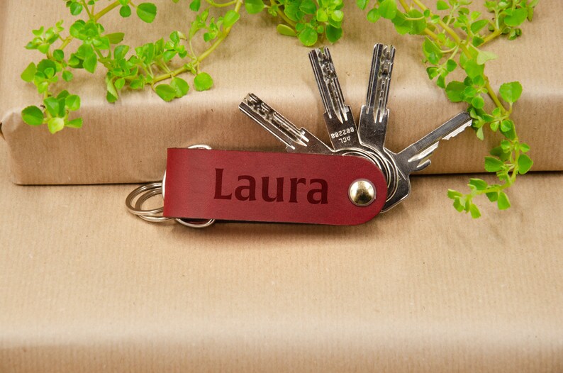 May include: Red leather keychain with the name "Laura" engraved on it. The keychain has a silver ring and is attached to a set of keys.