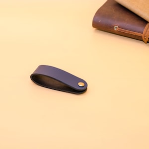 May include: A dark blue leather bookmark with a gold metal accent. The bookmark is on a light beige surface.