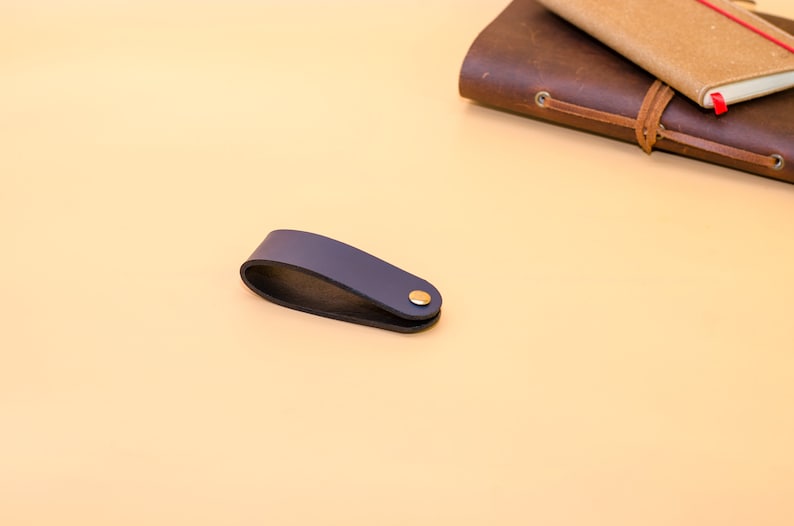 May include: A dark blue leather bookmark with a gold metal accent. The bookmark is on a light beige surface.