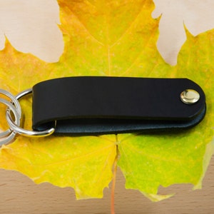 May include: A black leather key chain with a gold button and a silver key ring. The key chain is lying on a yellow maple leaf.