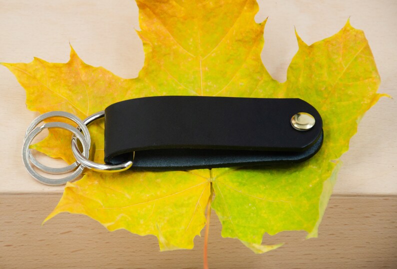May include: A black leather key chain with a gold button and a silver key ring. The key chain is lying on a yellow maple leaf.